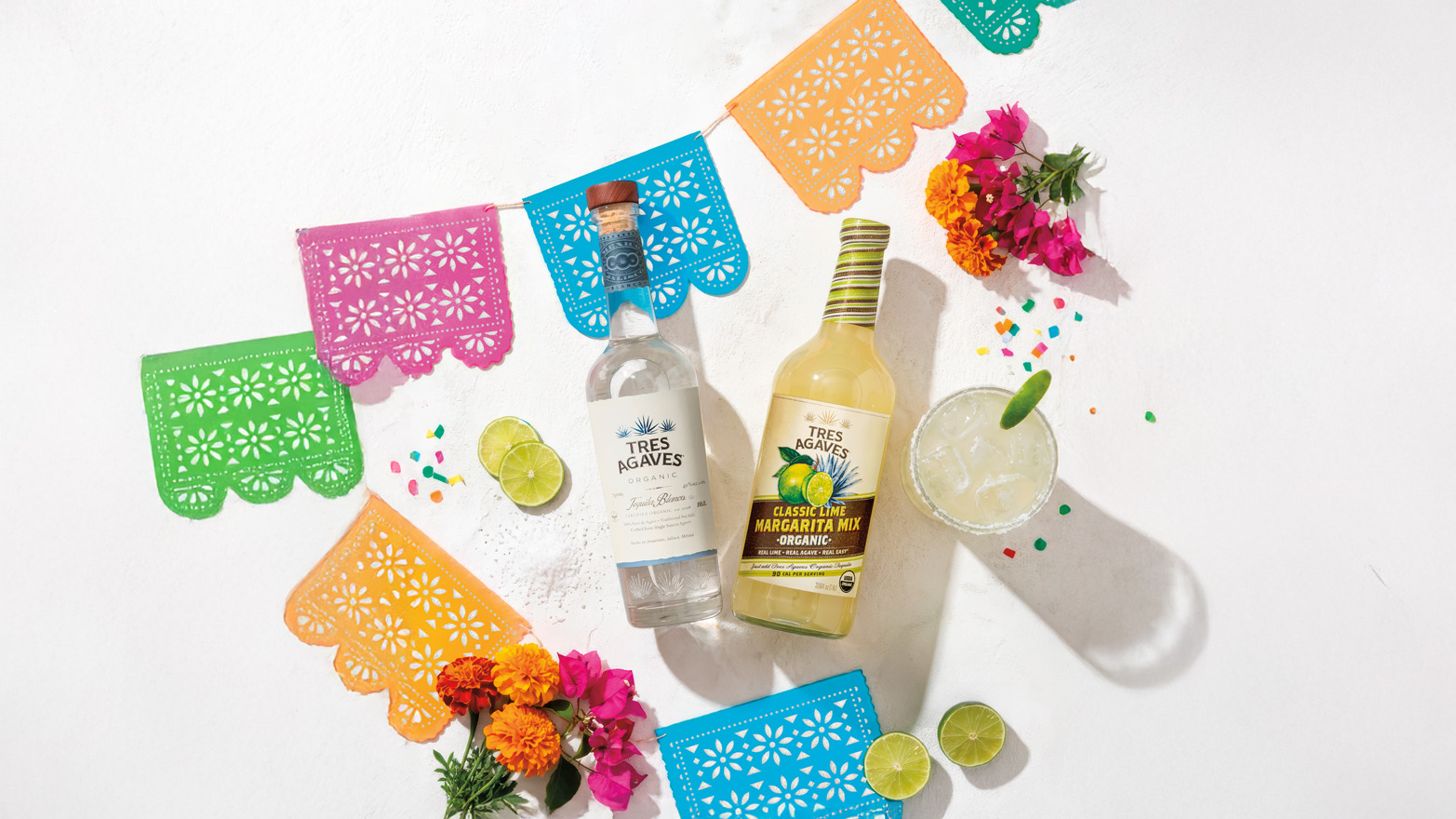 Festive Cinco de Mayo tabletop scene with bottles of Tres Agaves Organic Classic Lime Margarita Mix and Tres Agaves Organic Blanco, a mixed margarita drink in a glass, colorful flowers, and colorful stringed decorative flags